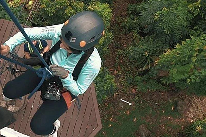 Eine Person mit Helm und Klettergurt, an einer Holzplattform in ein Seil eingehängt